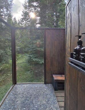A wooden cabin with a stone-tiled floor, a bench, and a shelf with two black bottles labeled 'SHAMPOO' and 'WASH'.