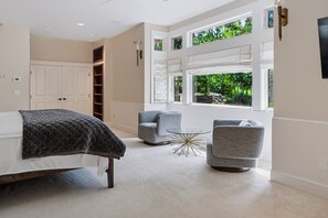 A bedroom with a bed, two armchairs, a small table, and a view of greenery through large windows. - Kai|Ridge At The Running Y (Klamath Falls)