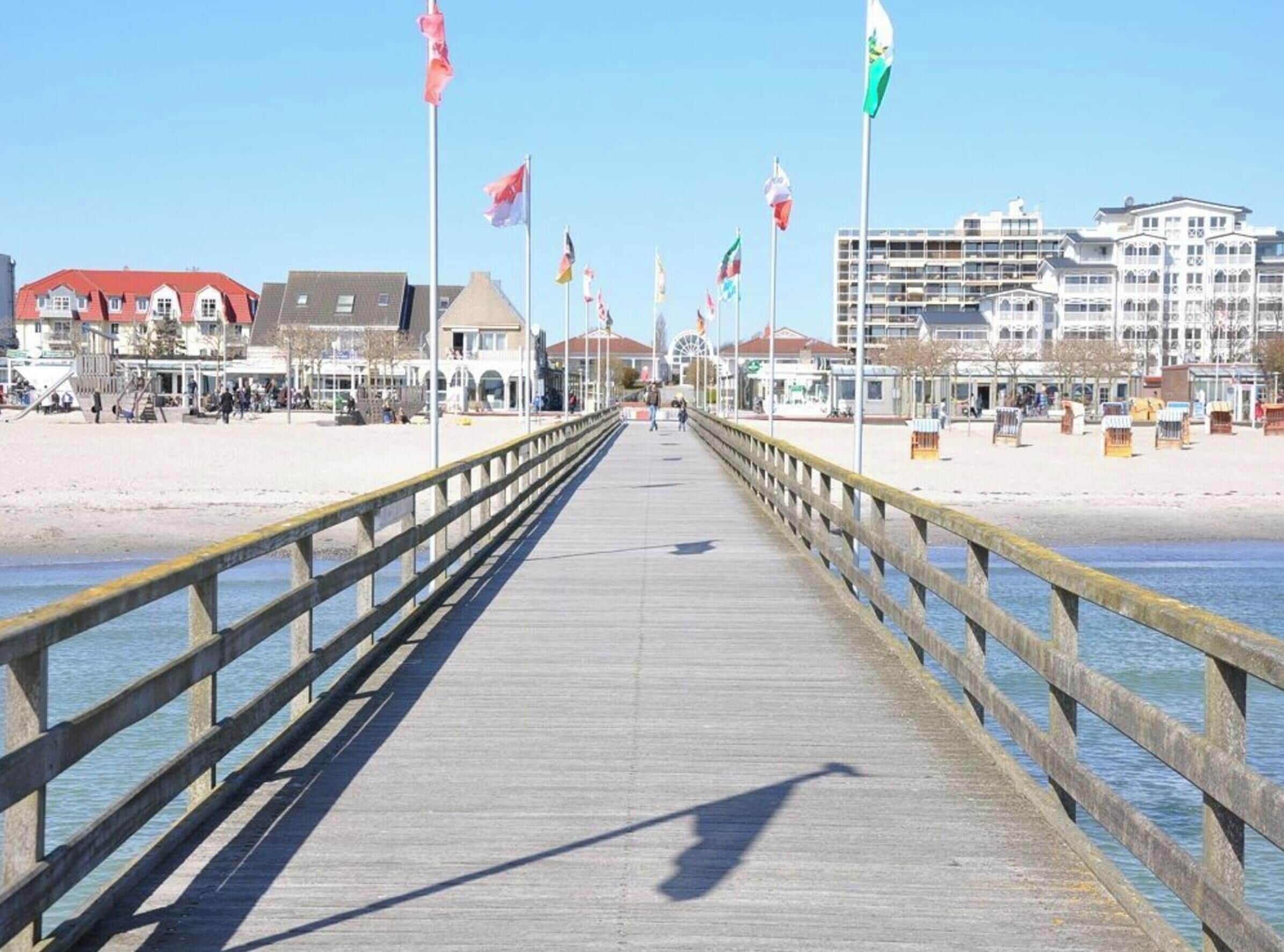 Eine Pier, die sich ins Wasser erstreckt, mit einer Holzreling, Flaggen und Gebäuden im Hintergrund.