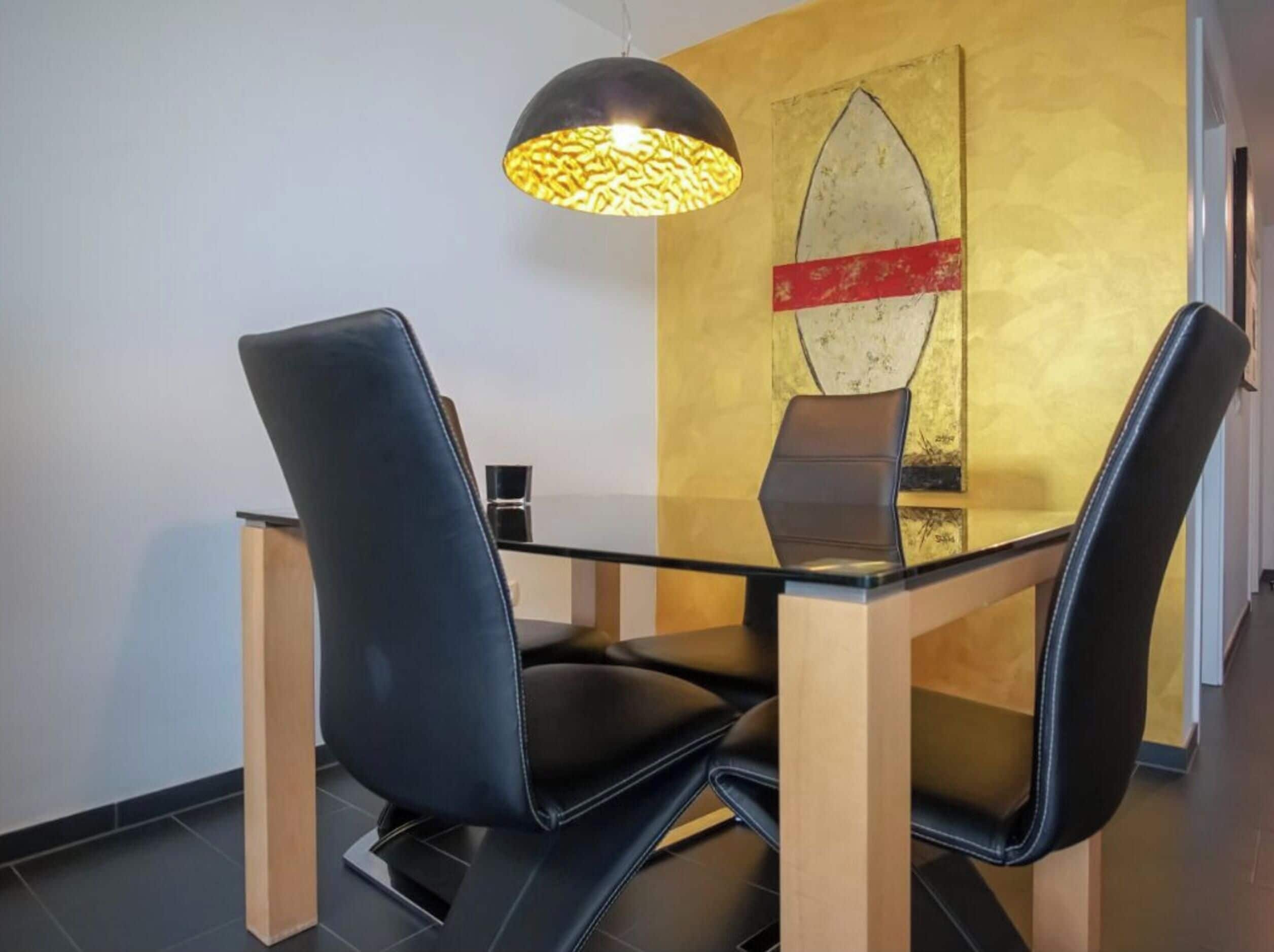 Un espace repas moderne avec une table en verre et des chaises en cuir noir, un luminaire suspendu et un mur décoré d’un motif de planche de surf.
