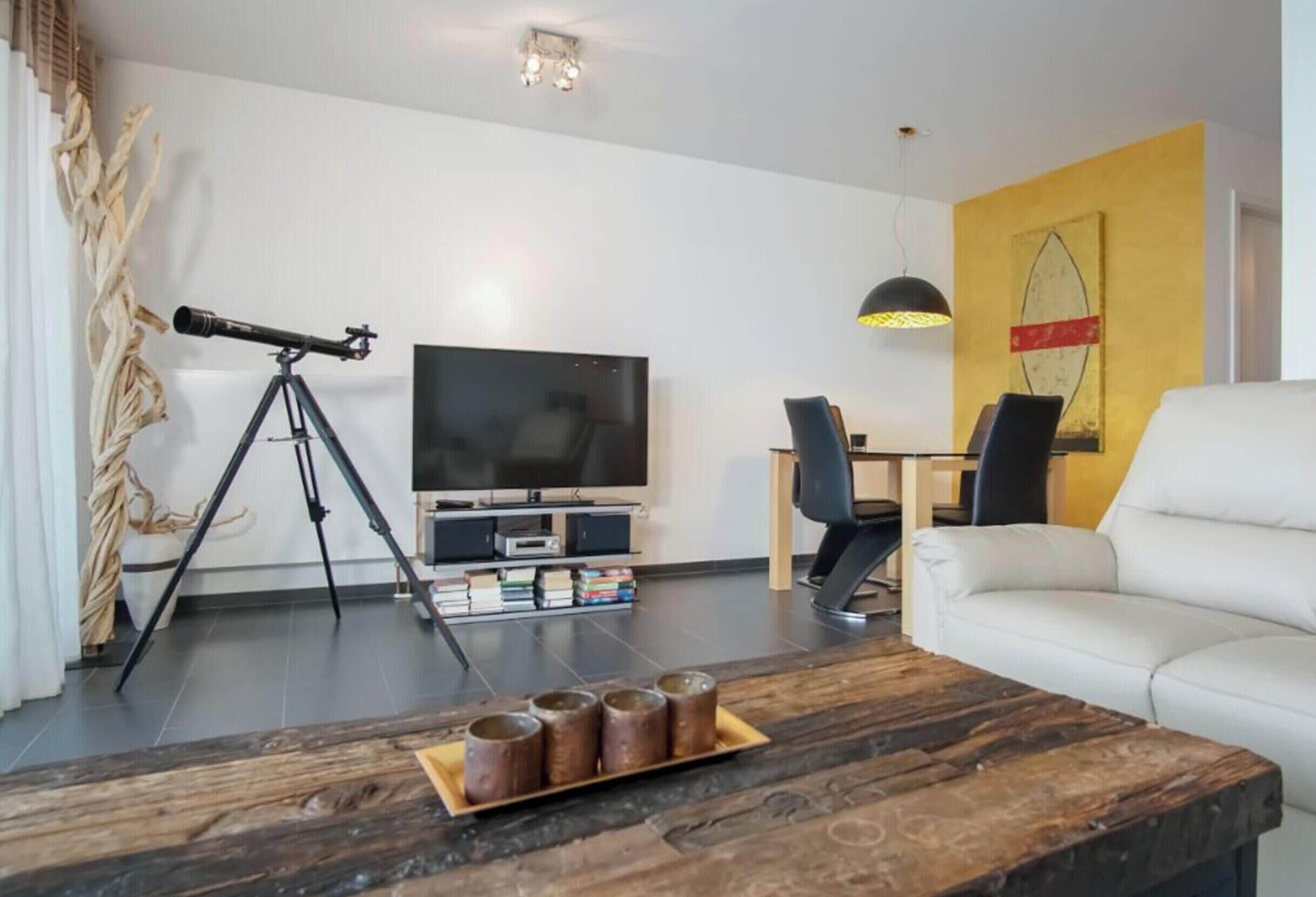 Una sala de estar con un telescopio, un televisor de pantalla plana, un sofá blanco y una mesa de centro de madera con libros.