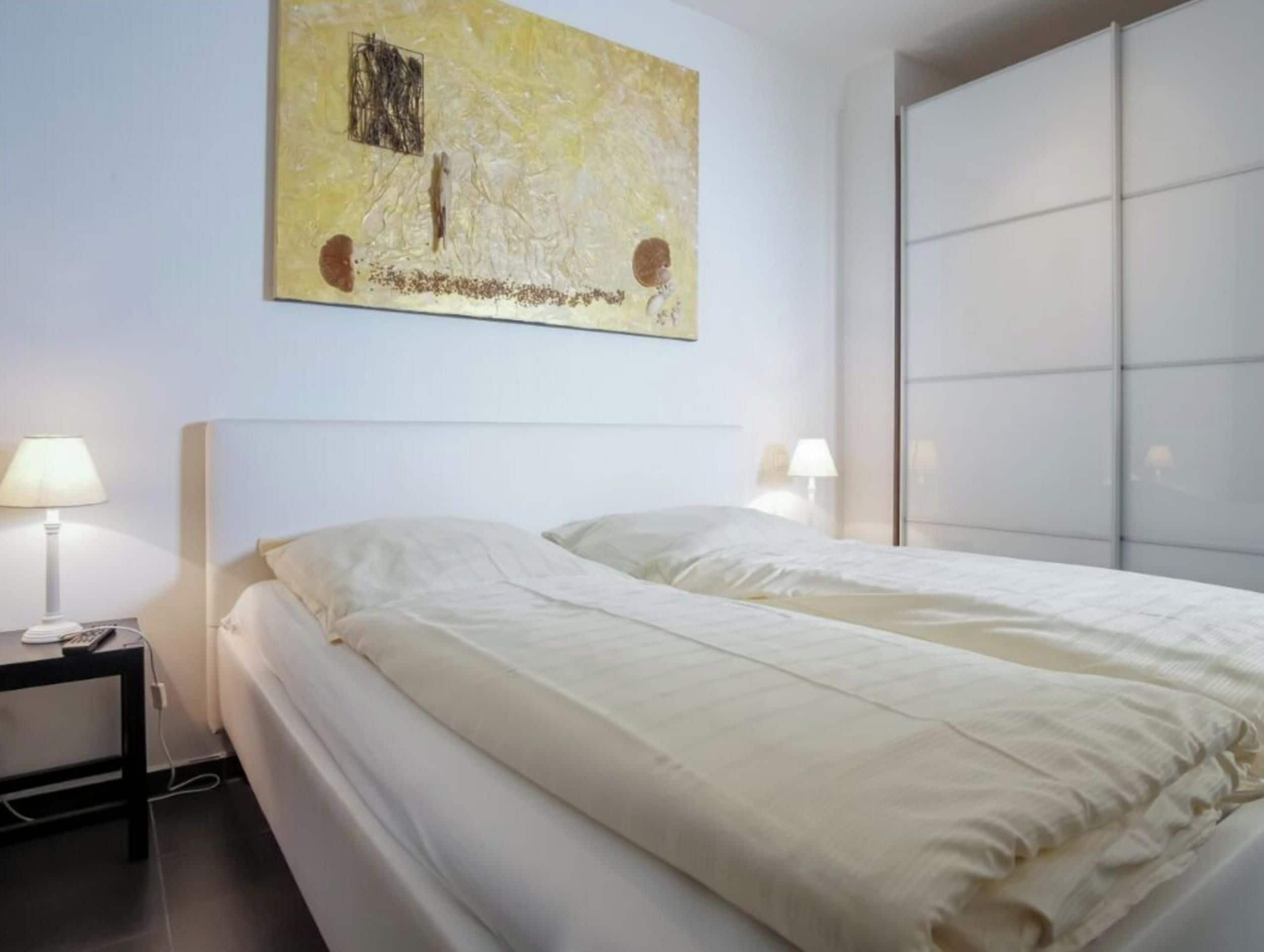 Quarto de hotel com cama, duas luminárias de cabeceira, uma obra de arte na parede e um armário com portas deslizantes.