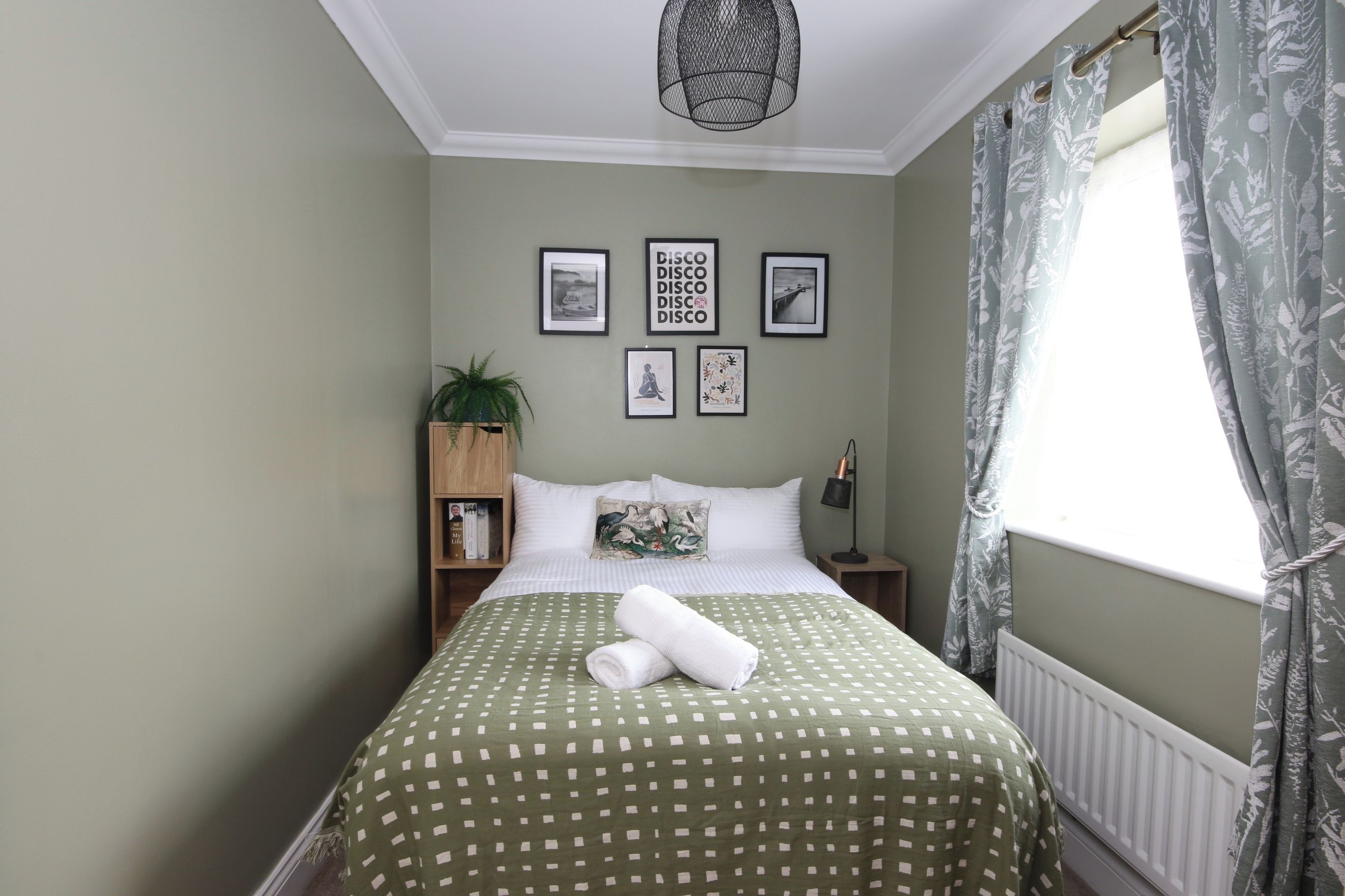 A bedroom with a bed, a nightstand, a lamp, a potted plant, and framed pictures on the wall.