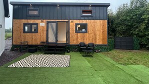 Ein modernes, kompaktes Gebäude mit Holzfassade, Glastür und einer kleinen Außenbereich-Sitzgelegenheit mit Stühlen und Tisch.