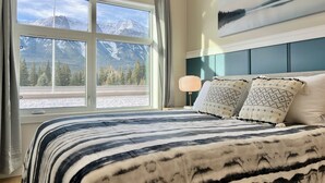 A bedroom with a large window offering a mountain view, a bed with patterned bedding, and a painting on the wall. - Separate-Address Combo • Hot Tub • Parking • Air Conditioner, Sleeps up to 17 (Harvie Heights)