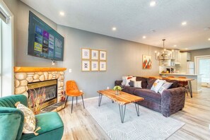 A modern living room with a stone fireplace, a flat-screen TV mounted on the wall, a sofa, a coffee table, and a dining area with a kitchen in the background. - Separate-Address Combo • Hot Tub • Parking • Air Conditioner, Sleeps up to 17 (Harvie Heights)