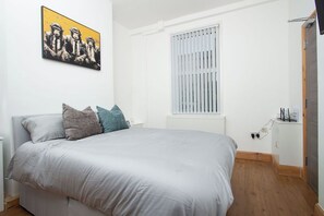 Ein Schlafzimmer mit einem Bett, Kissen, einem Heizkörper, einem Fenster mit Jalousien und einem kleinen Schrank.