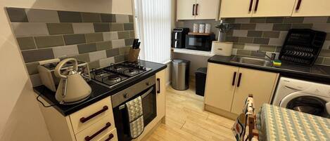 A compact kitchen with a gas stove, oven, and microwave. There is a sink with a dish rack, and a washing machine is visible in the background.