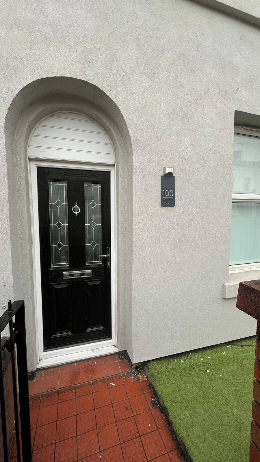 Eine schwarze Tür mit Fensterglas und Metallbeschlägen, ein weiß gefasster Bogen und ein kleines Schild mit der Nummer '100' an der Wand.