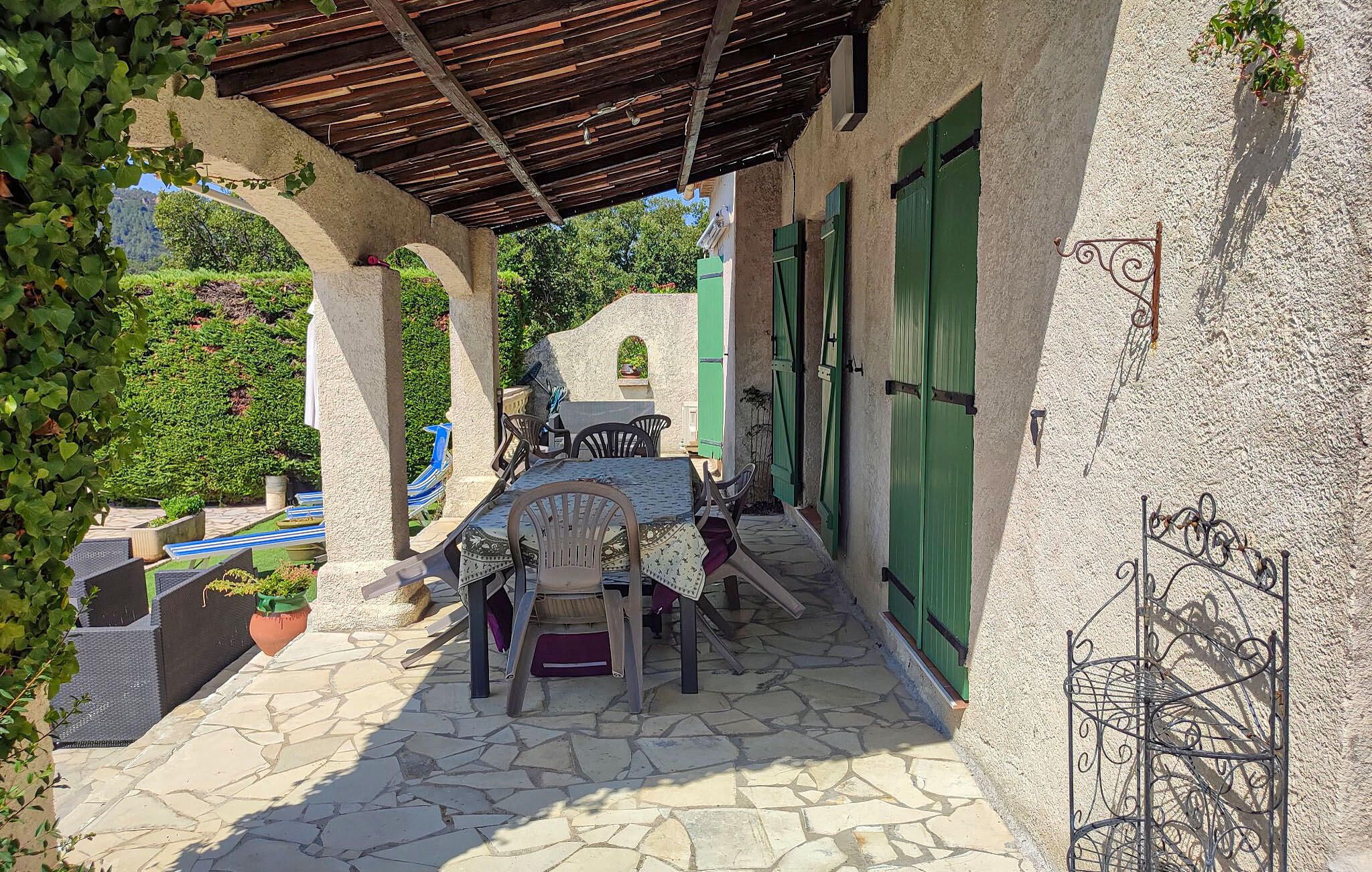 Une terrasse extérieure couverte, dotée d’un sol en pierre, d’une table avec des chaises et de volets verts aux fenêtres.