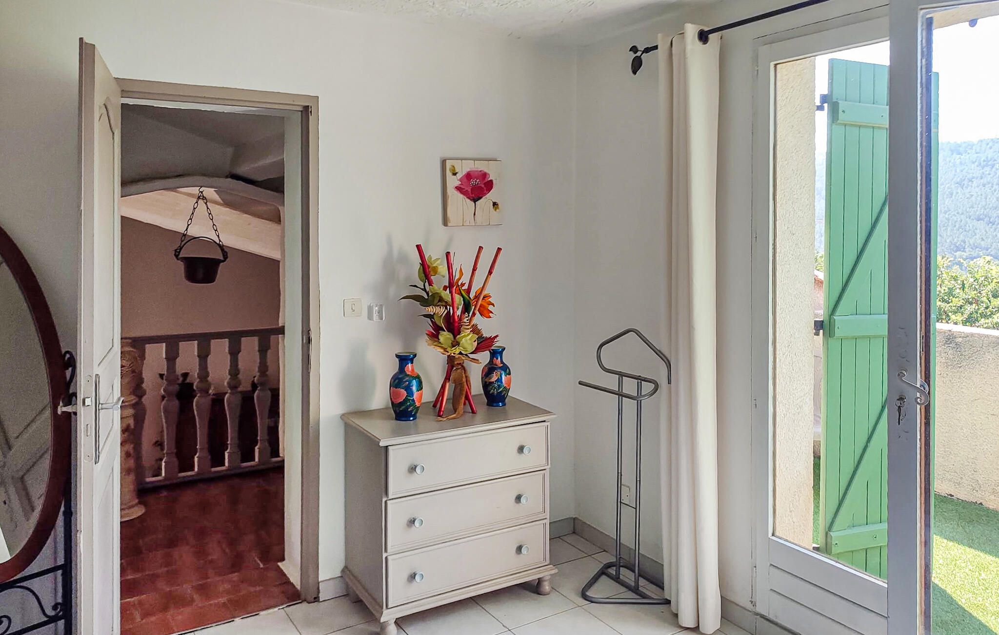 Une pièce avec une commode blanche, un vase rempli de fleurs, un tableau accroché au mur et une vue sur la verdure à l’extérieur.