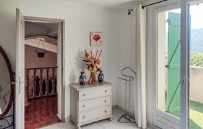 A room with a white dresser, a vase with flowers, a picture on the wall, and a view of greenery outside.