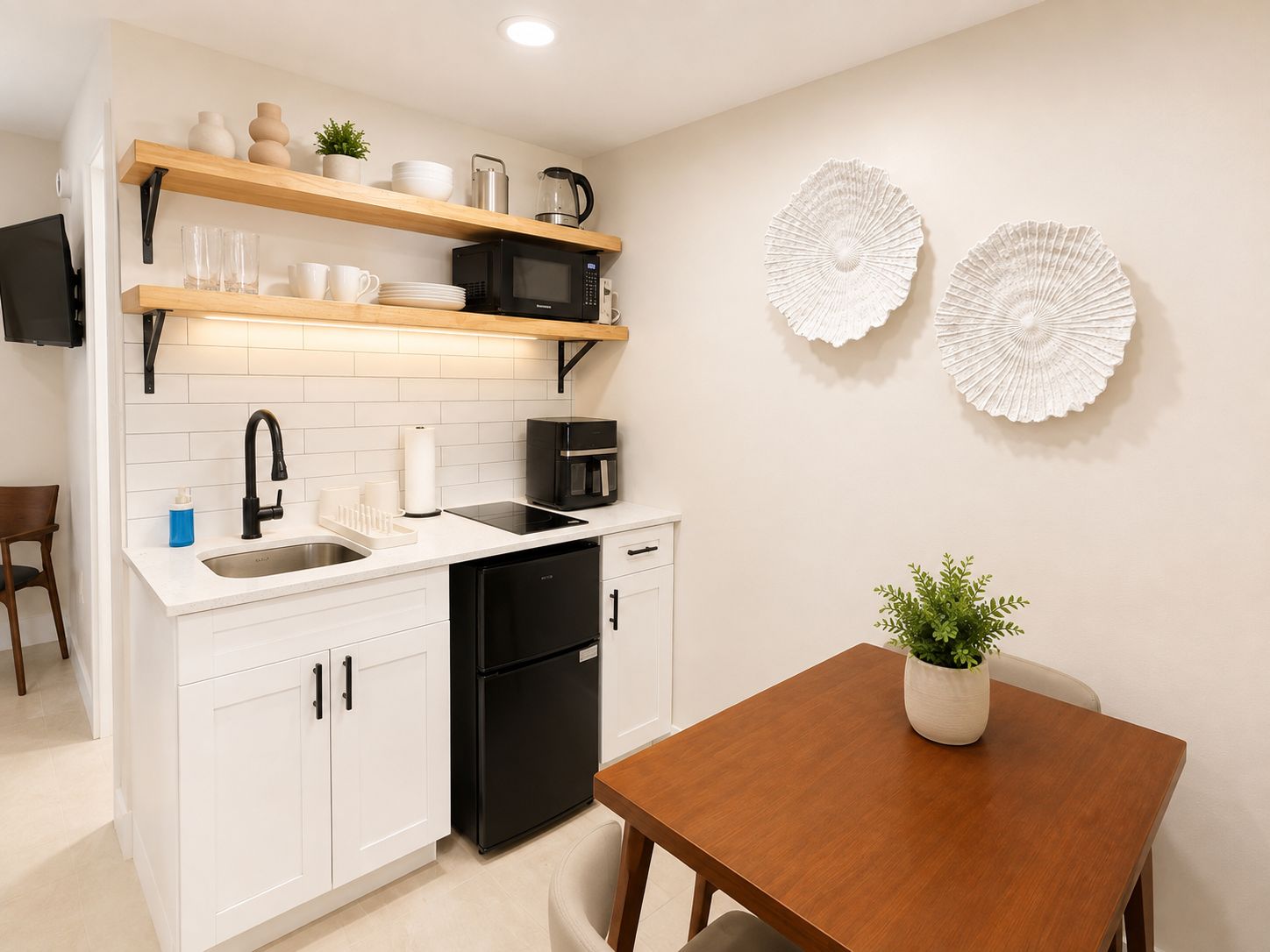 Cocina compacta con armarios blancos, lavavajillas negro, cafetera y microondas. Hay dos adornos blancos en la pared y una planta en maceta sobre la mesa de comedor.