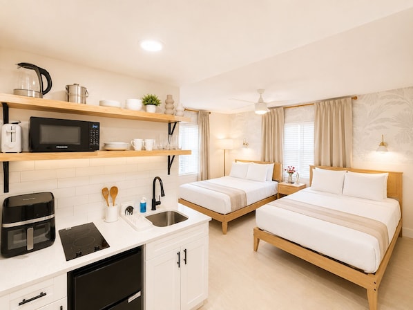 A hotel room with a kitchenette, two beds, a ceiling fan, and a window with curtains.