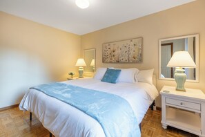A hotel room with a large bed, two bedside tables with lamps, a wall-mounted mirror, and a floral artwork above the bed. - Whispering Pines- 5Br Wooded Retreat in Cary (Cary)