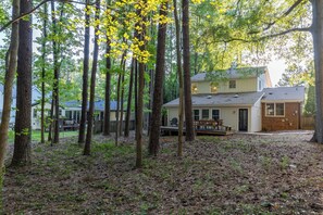 A two-story house surrounded by tall trees with a deck and outdoor seating area. - Whispering Pines- 5Br Wooded Retreat in Cary (Cary)