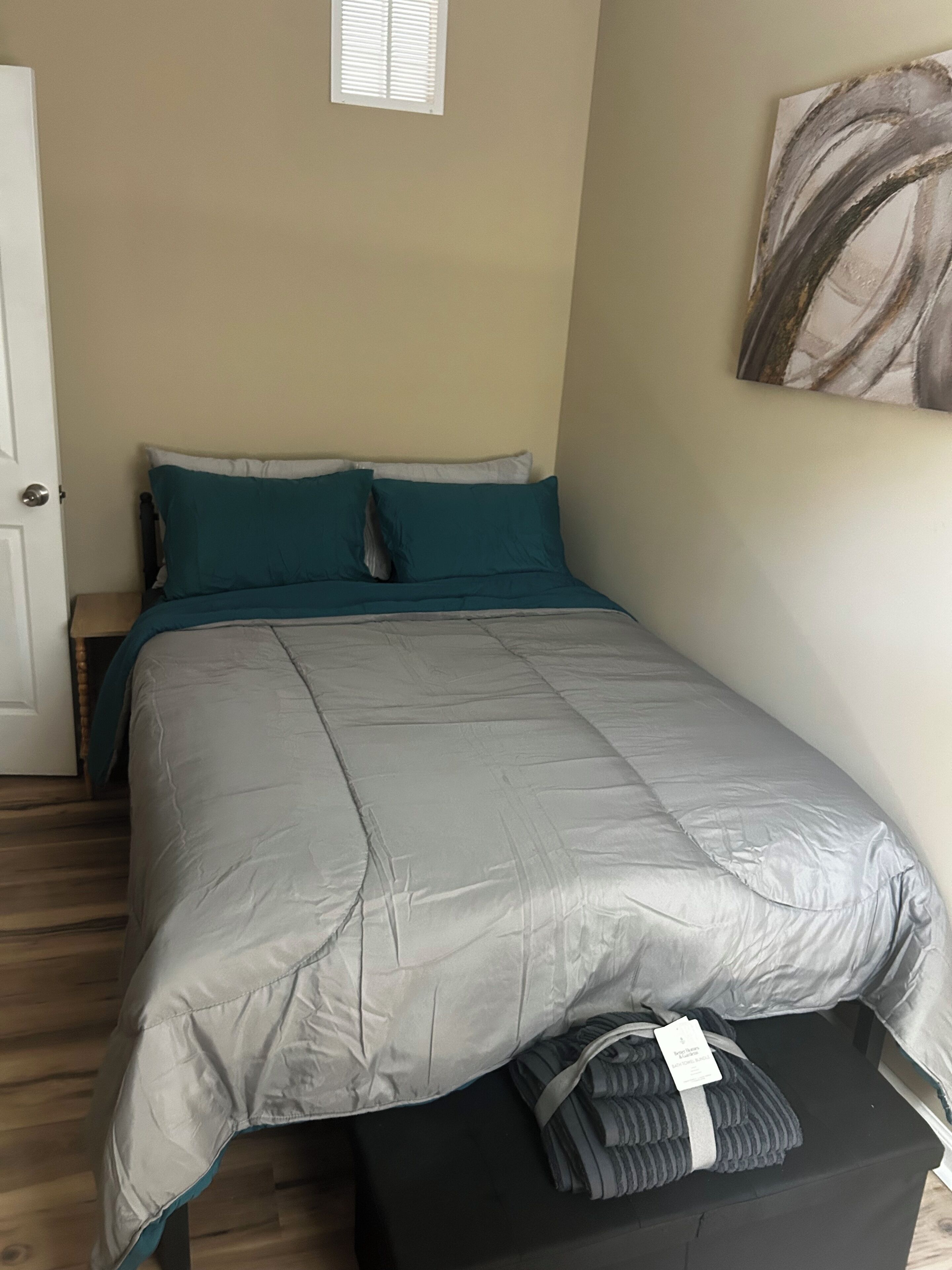 Un dormitorio con cama, almohadas color turquesa, edredón gris, mesita de noche negra, ventana con persianas y un cuadro en la pared.