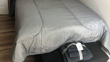 Un dormitorio con cama, almohadas color turquesa, edredón gris, mesita de noche negra, ventana con persianas y un cuadro en la pared.