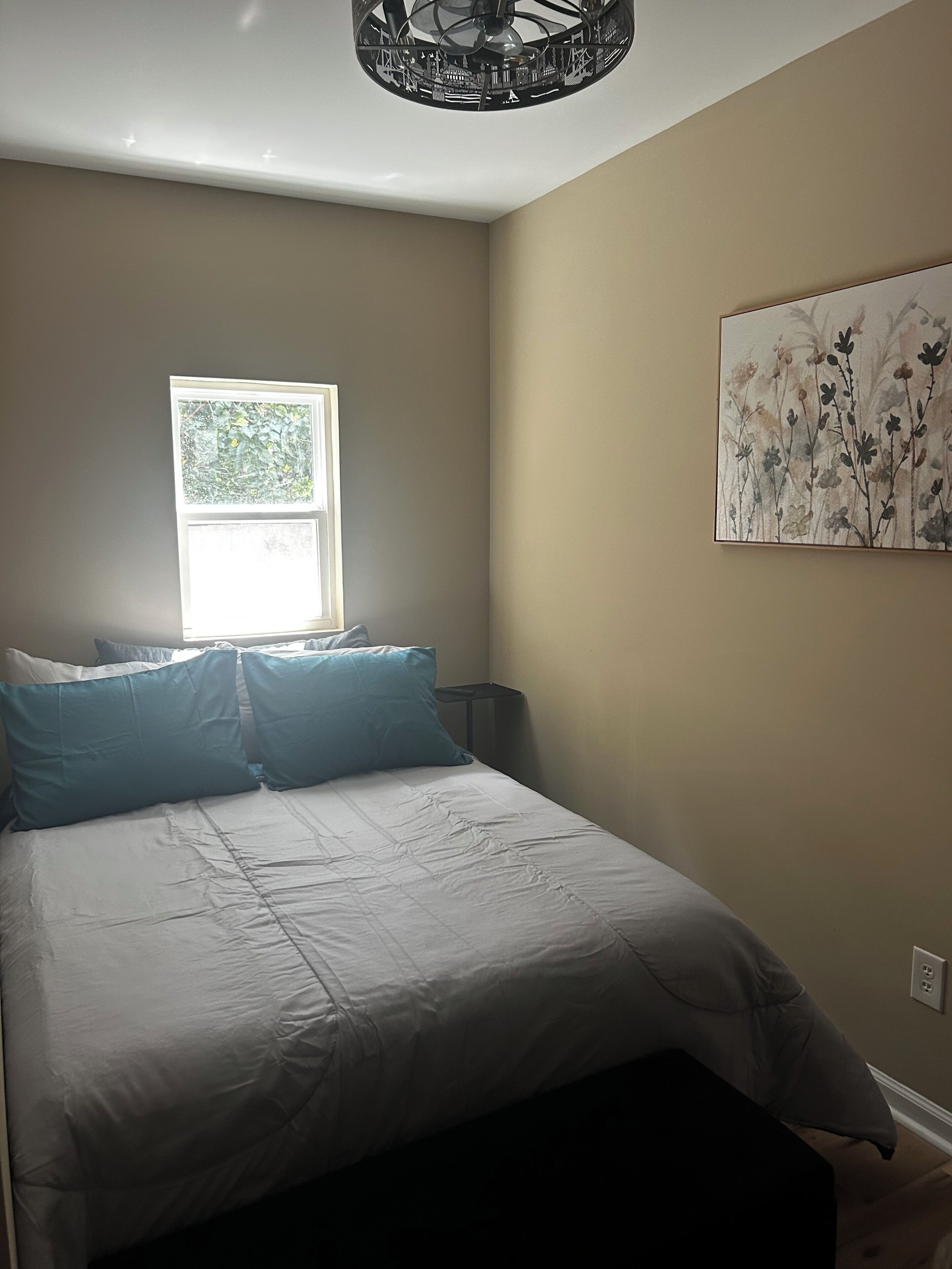 Un dormitorio con cama, almohadas, una ventana, un cuadro colgado en la pared y una lámpara en el techo.