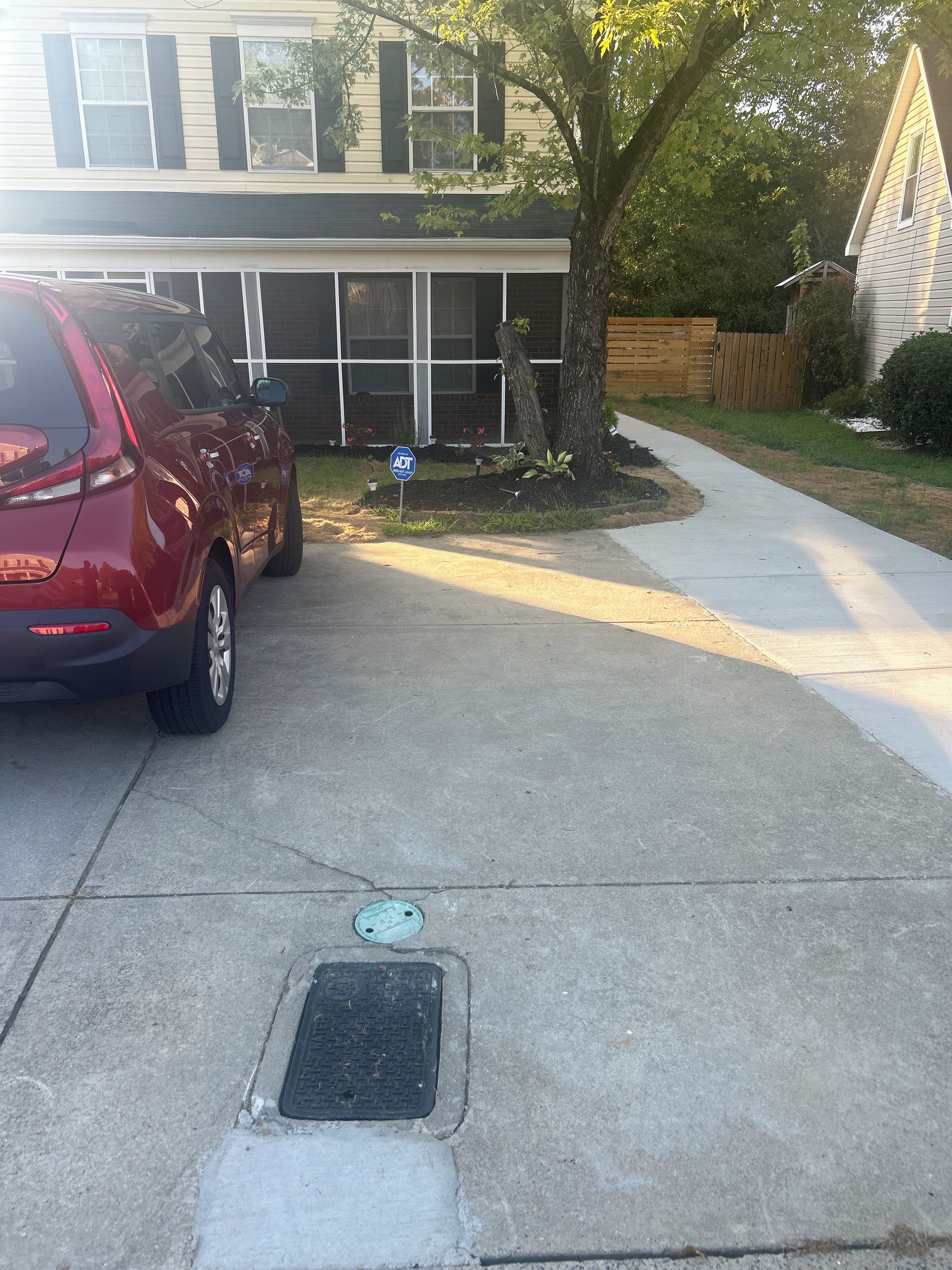 Un camino de entrada residencial con un coche aparcado, un árbol y una acera que lleva a una casa.