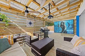 An outdoor deck with wicker furniture, a grill, and a mounted TV screen. - Tremendous 2-bedroom house with AC in Panama City Beach (Panama City Beach)