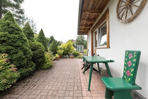A paved courtyard with a wooden bench and a table, surrounded by greenery and a building with a wooden wheel. - Gronik Mountain Resort 1 Apartment (Kościelisko)