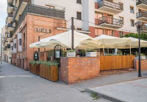 Un edificio de ladrillo con mesas al aire libre y sombrillas, un letrero que dice 'BOSKA' y una cerca de madera con plantas.