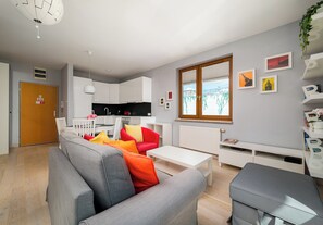 Una sala moderna con un sofá gris, un sillón rojo y una mesa de centro blanca. Se ve una cocina con armarios blancos y una encimera negra al fondo.