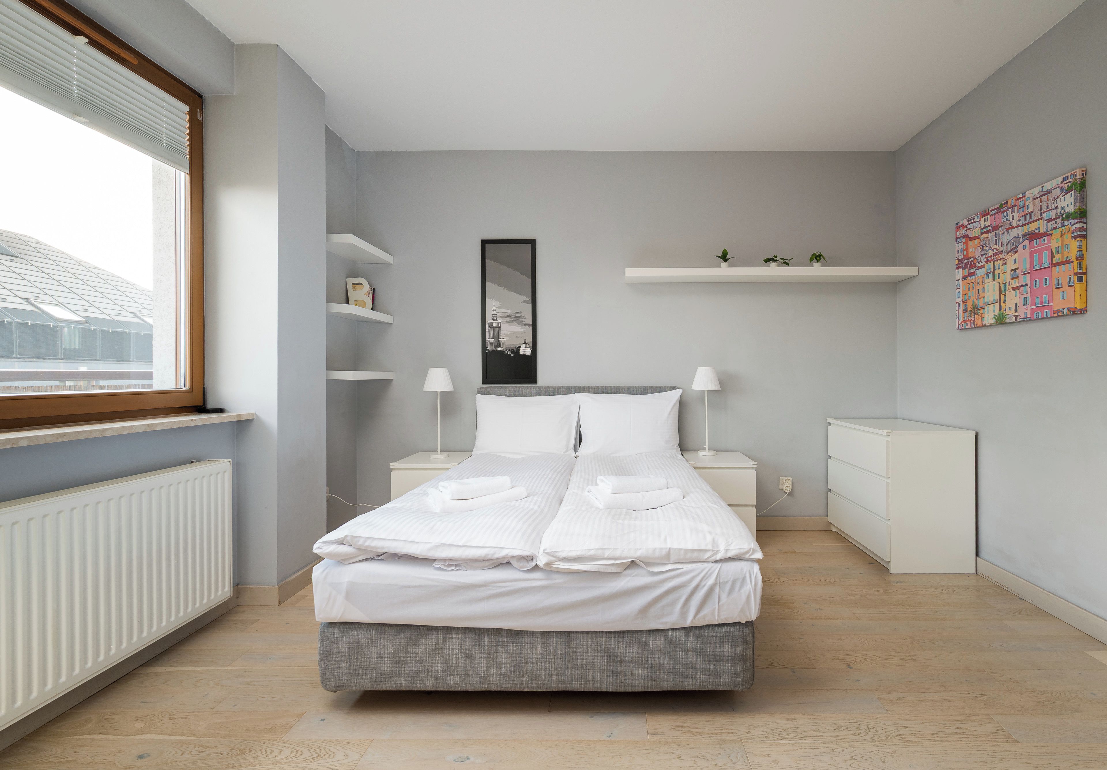 Une chambre moderne avec un lit, des tables de chevet, un radiateur, une fenêtre et une vue sur le paysage urbain.