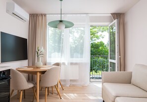 A modern living room with a dining area, a flat-screen TV, a sofa, and a balcony with a view of trees.