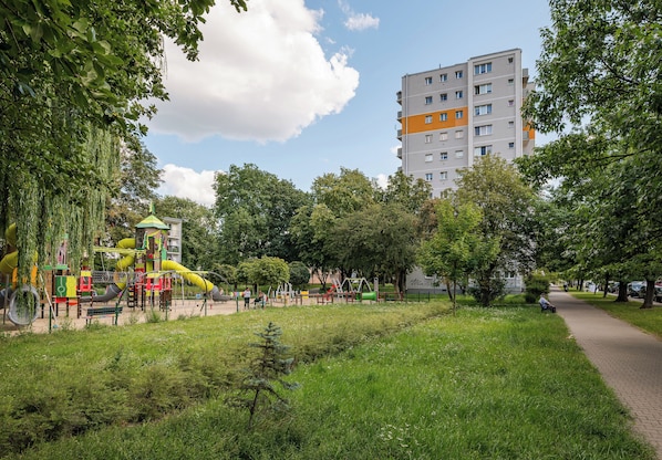 A park with playground equipment, a walking path, and a multi-story building in the background.