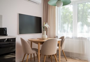 A modern dining area with a round table, four chairs, a vase with flowers, a large flat-screen TV, and a window with a view of trees. - Kinowa Deluxe | Warsaw Arena Vibes (Warszawa)