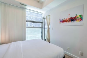 A hotel room with a bed, a large window with curtains, and a cityscape painting on the wall.