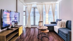 Ein modernes Wohnzimmer mit einem Flachbildfernseher, einem Sofa, einem kleinen runden Tisch und Blick auf die Stadt durch große Fenster.