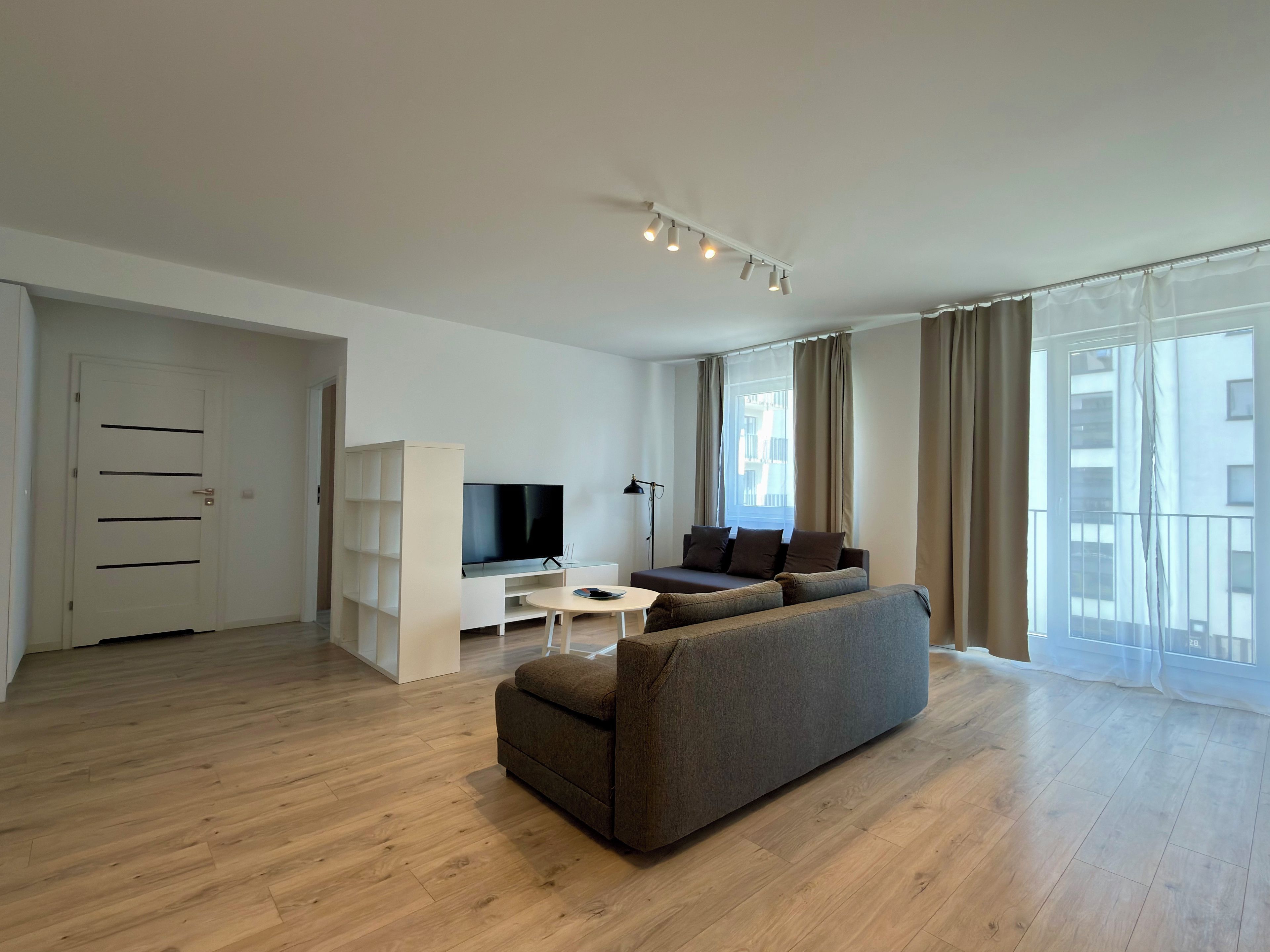 Ein modernes Wohnzimmer mit einer Couch, einem Fernseher, einem weißen Schrank und einem großen Fenster mit Vorhängen.