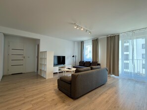 Un salon moderne avec un canapé, un téléviseur, un meuble blanc et une grande fenêtre avec des rideaux.