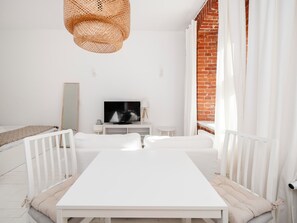 A modern, minimalist living room with a white sofa, a dining table, a television, and a wicker ceiling light. - Park Źródliska White & Brick Studio (Łódź)