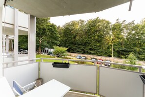 Ein Balkon mit einem weißen Tisch und Stuhl, einer hängenden Blumenampel und Blick auf einen Parkplatz mit Autos und Bäumen.