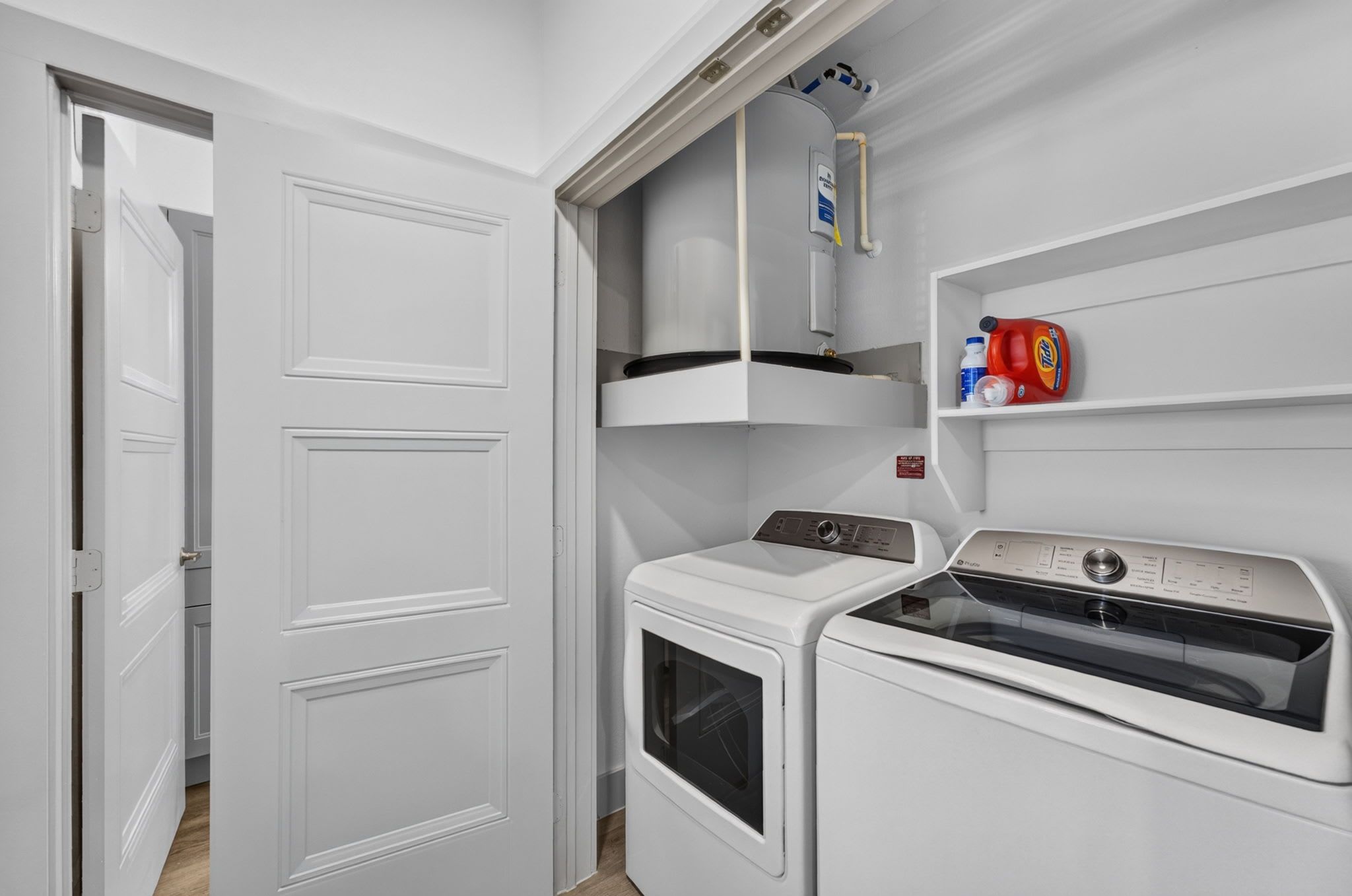 A compact laundry room with a washing machine, dryer, and a shelf with detergent bottles.
