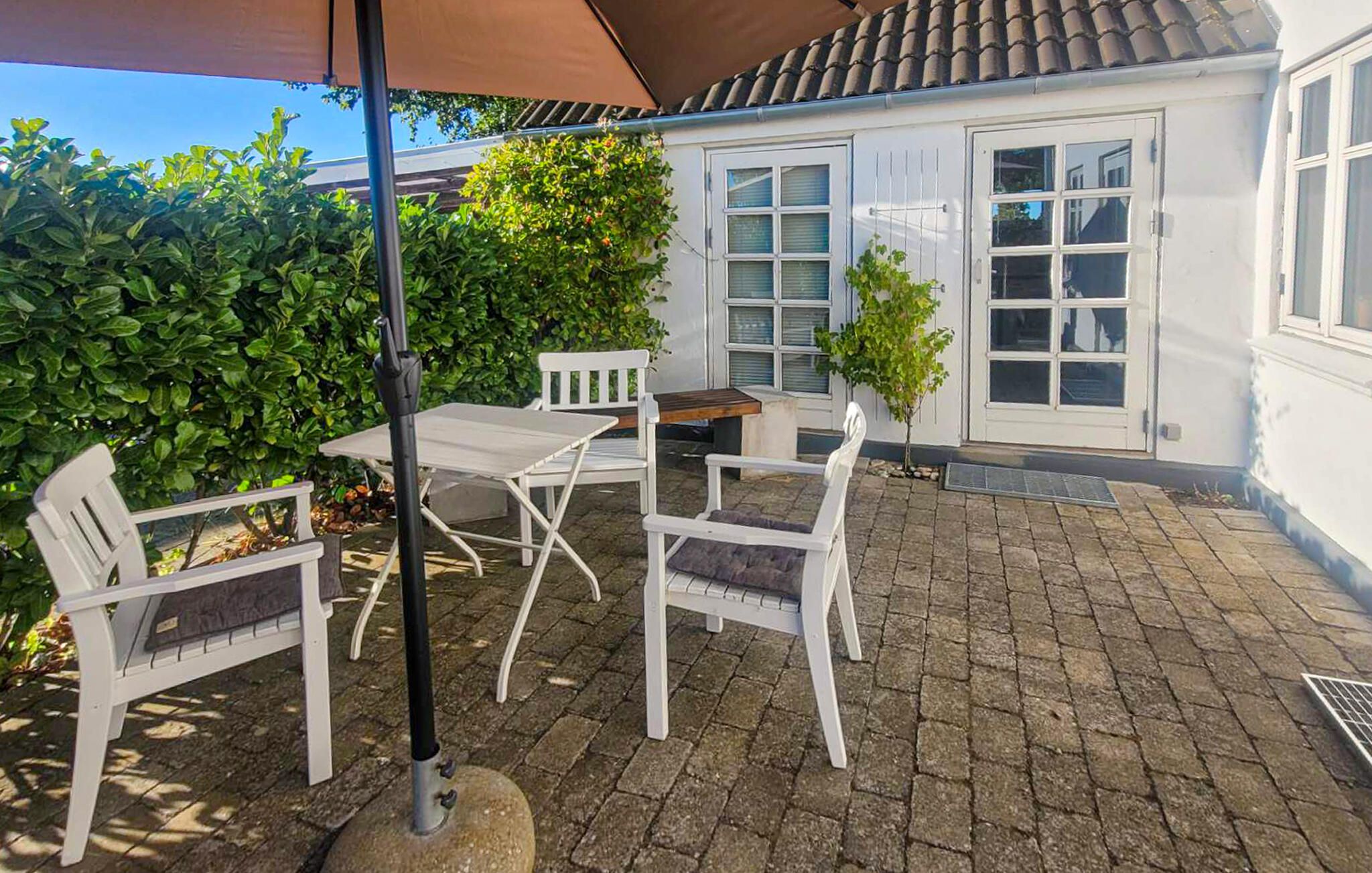 An outdoor patio with a table and chairs, a small tree, and a white building with windows.