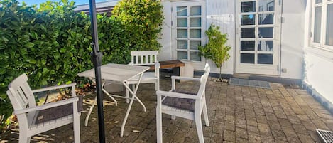 Un patio al aire libre con una mesa, sillas, un pequeño árbol y un edificio blanco con ventanas.