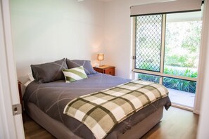 A bedroom with a bed, a nightstand, a lamp, and a view through a window. - Hop, Skip and a Jump - Entire vacation home on acreage (Landsborough)