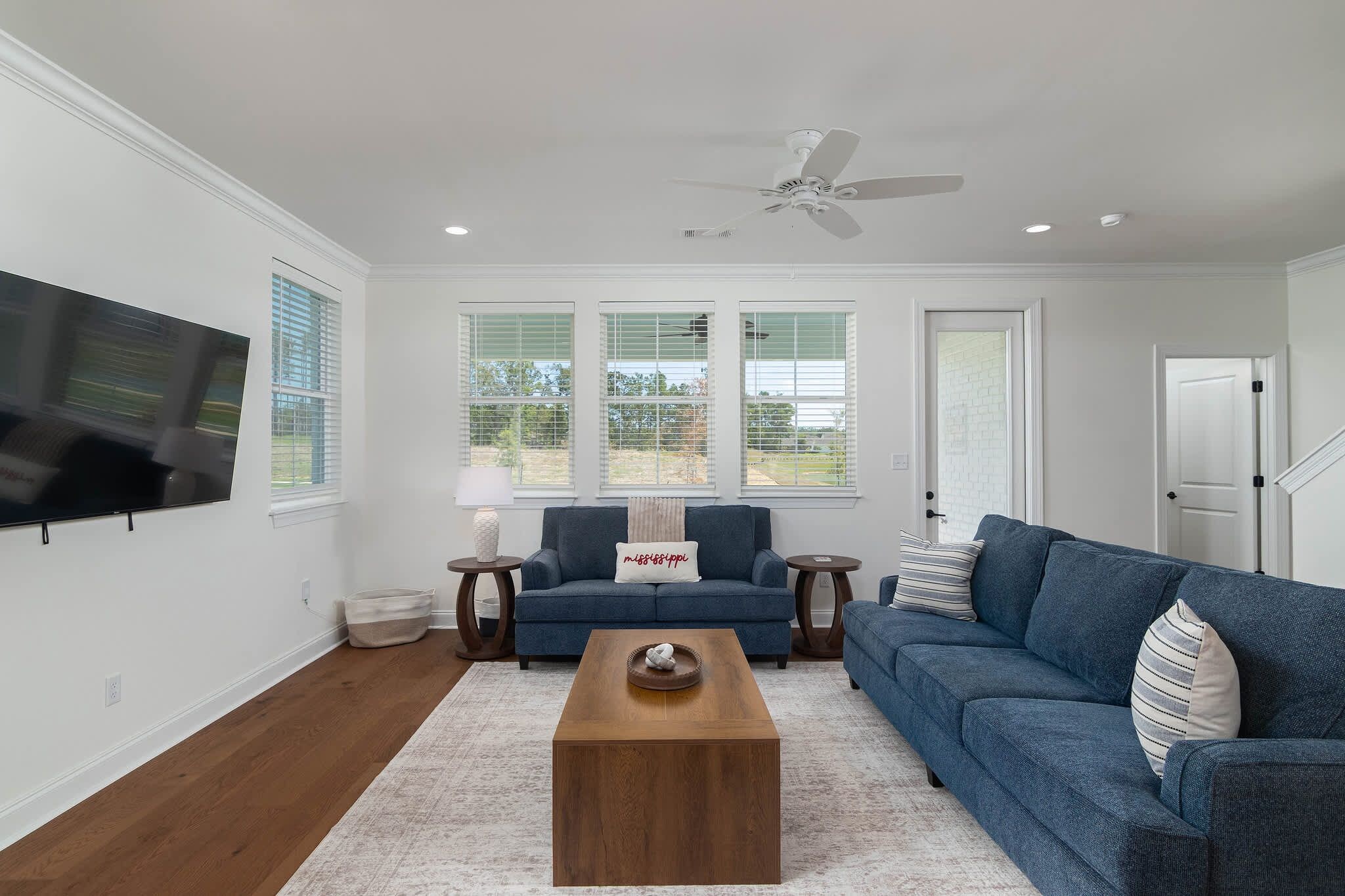 A living room with a flat-screen TV, a blue sofa, a wooden coffee table, and a ceiling fan.