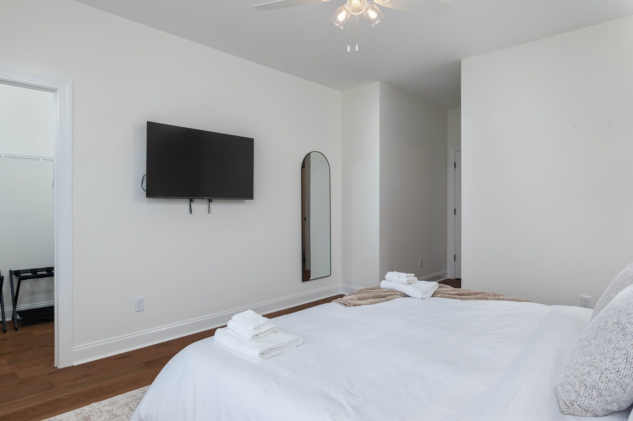 A bedroom with a bed, a TV mounted on the wall, a mirror, and a door leading to another room.