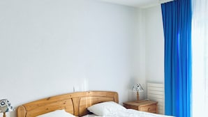 A single-bed room with a wooden headboard, white bedding, a bedside lamp, a nightstand, and a window with blue curtains. - Haus Alpina (CharmingStay). 2.5-Zimmerwohnung mit zwei Balkonen by Interhome (Mols)