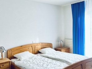 Habitación con una cama individual, cabecera de madera, ropa de cama blanca, lámpara de noche, mesita de noche y una ventana con cortinas azules.
