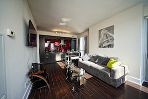 A modern living room with a grey sofa, a glass coffee table, and a dining area in the background.