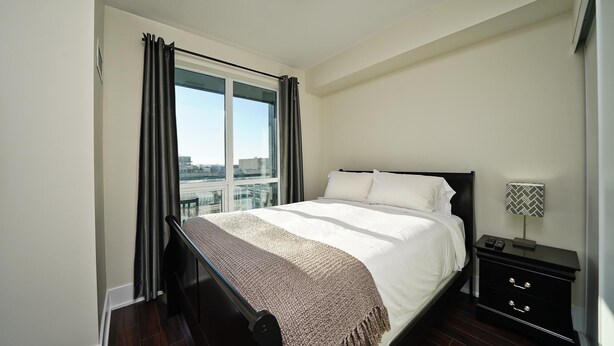 Ein Schlafzimmer mit einem großen Bett, einem Nachttisch mit Lampe, einem Fenster mit Vorhängen und Blick auf die Stadt.
