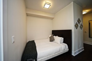 A small, neatly made bed with white bedding and a dark headboard in a minimalistic room with white walls and a dark floor.