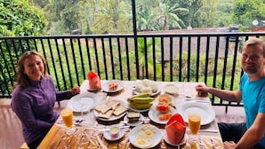 Mesa de comedor con diversos platos y bebidas, rodeada por una barandilla con vistas a un paisaje verde y exuberante.